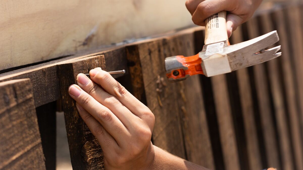 Close-up of hands driving a nail into a dark wood fence board with an orange-handled hammer